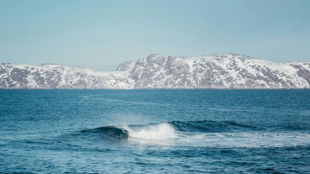 Captivating view of ocean waves crashing against a backdrop of snow-covered mountains under a clear sky.